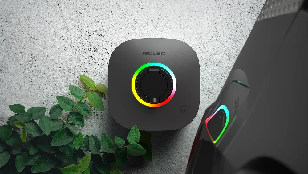 Wall-mounted Rolec EVO EV charger in dark grey, featuring a circular LED light ring displaying a multi-colour glow (green, yellow, red), installed against a light textured exterior wall with green foliage nearby. A second charger or vehicle with reflected LED glow is partially visible to the right.